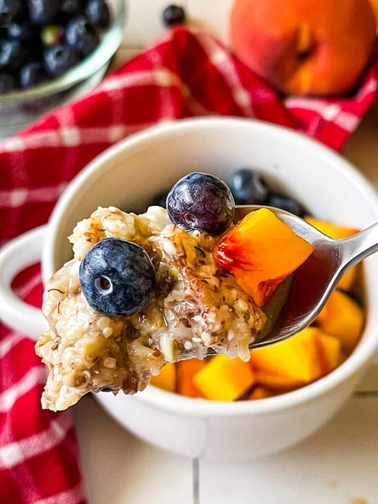 Oatmeal in a white bowl, with a spoon loaded with oatmeal blueberries and a piece of peach.