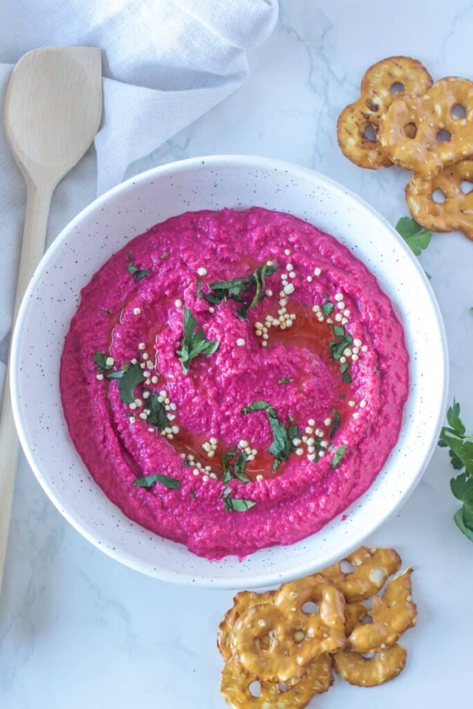 Pink beet hummus presented in a white bowl topped with green herbs beside a spoon and flat pretzels.