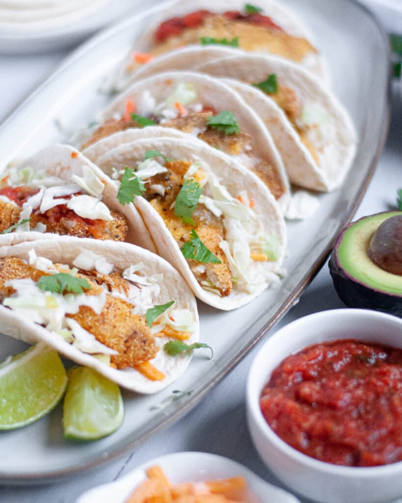 Soft shell small fish tacos lined up on an oval plate beside sliced lime and small bowls of salsa, cheese and avocado.