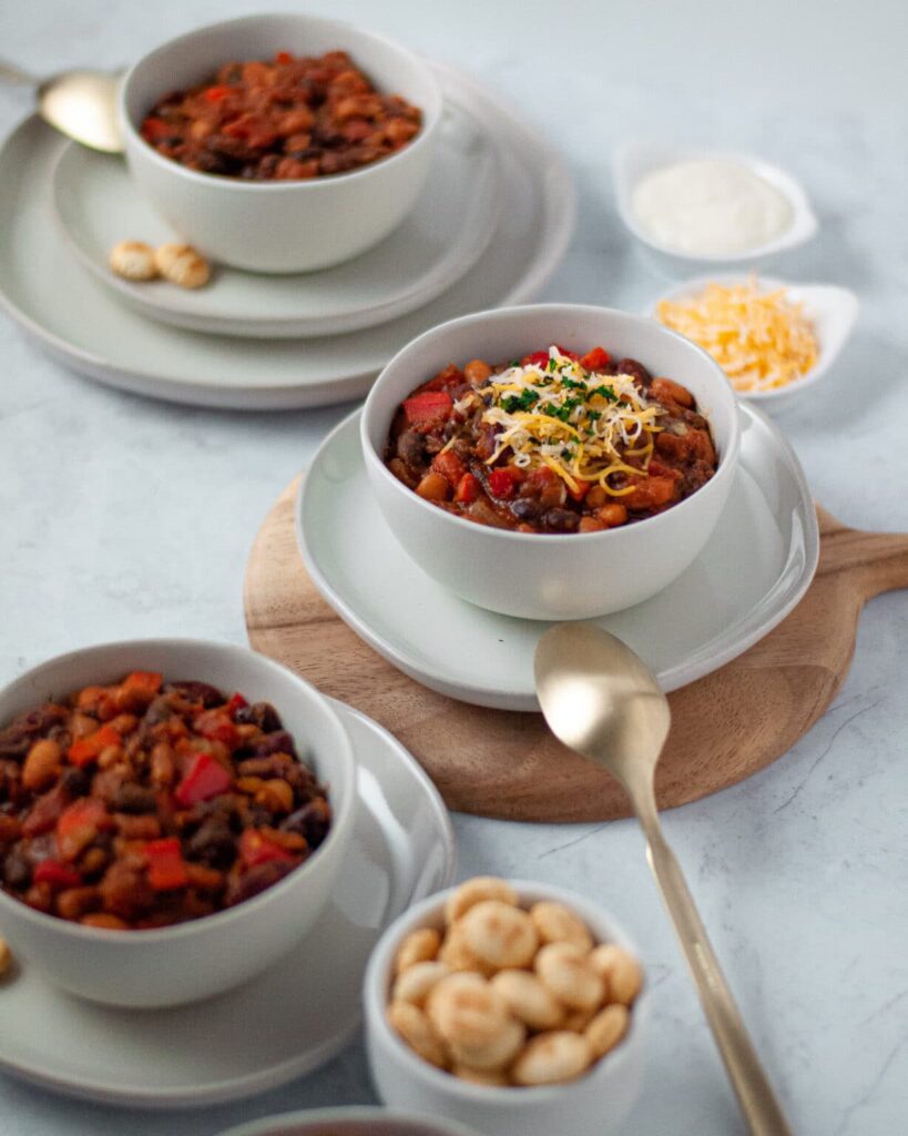 Three white bowls of chili on white plates topped with cheese and sour cream.