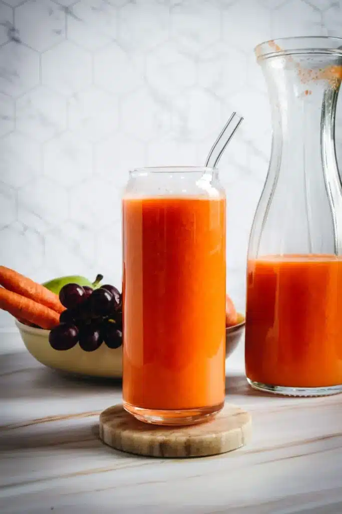 Large glass pitcher in background full of orange juice with a tall glass of orange juice front and center with a clear straw.