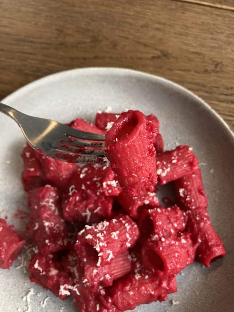 Heart healthy mac and cheese recipe that includes beet to make these noodles pink. Sprinkled with white cheese presented on a white plate with a fork lifting up one pink noodle.