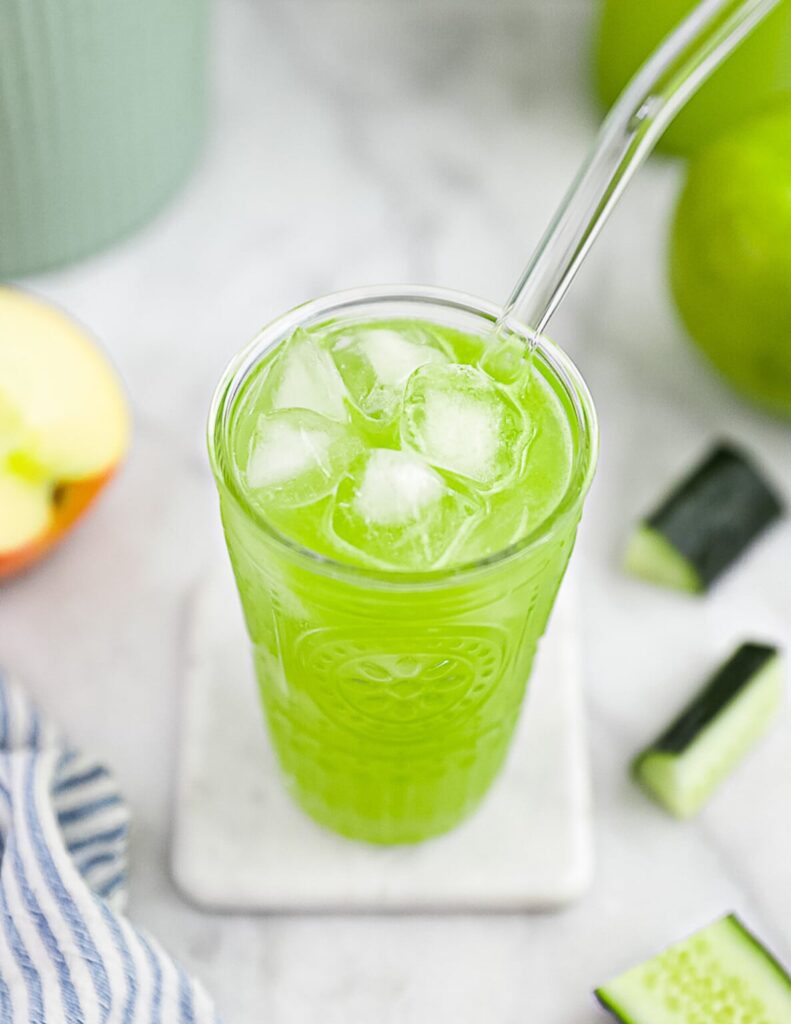 Green juice with ice in a clear glass with a clear straw on a white napkin beside sliced cucumber and a halved apple.