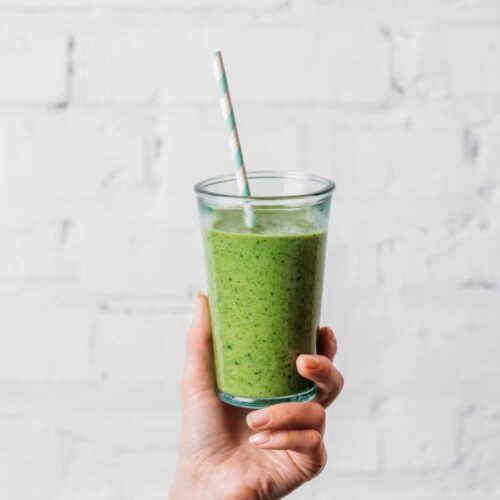 Green smoothie in a clear container being held by a hand with a green and white straw in front of a white background.