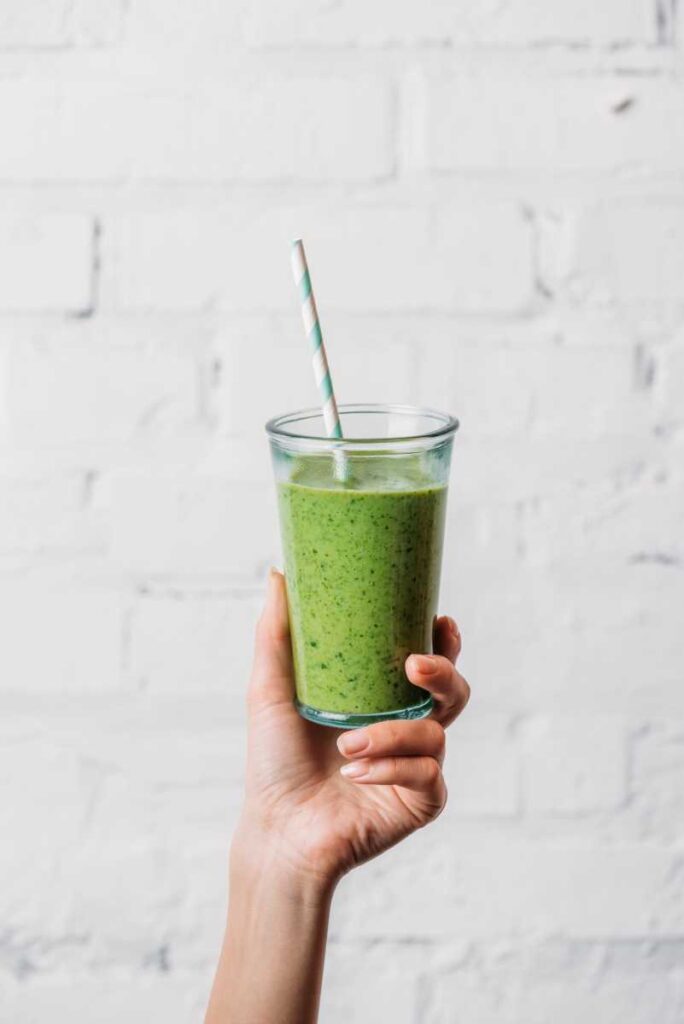 Green smoothie in a clear container being held by a hand with a green and white straw in front of a white background.
