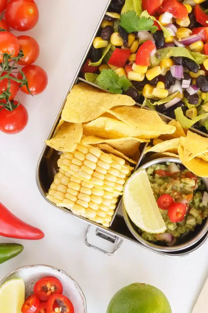 A low sodium lunch bento box style with a slice of corn on the cob, tortilla chips, dip and a black bean and corn salad. 