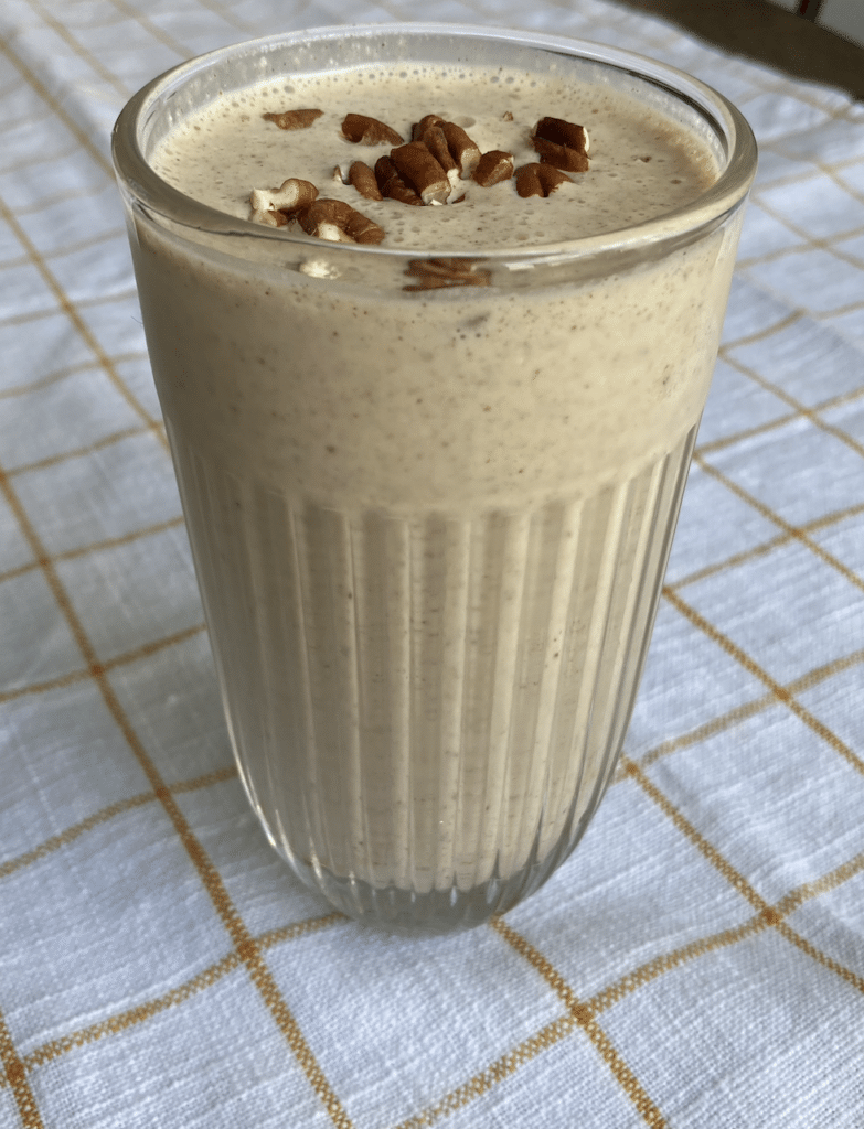 Pecan pie smoothie in a clear glass on a white and brown checkered tablecloth, topped with pecans.