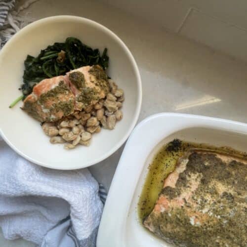 Cooked salmon in casserole dish beside a bowl with cooked spinach, beans and pesto butter salmon, beside a blue and white tea towel.