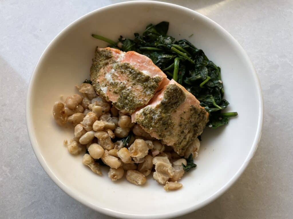 Cooked pesto butter salmon on top of cooked greens and white kidney beans in a white bowl on top of a white countertop.