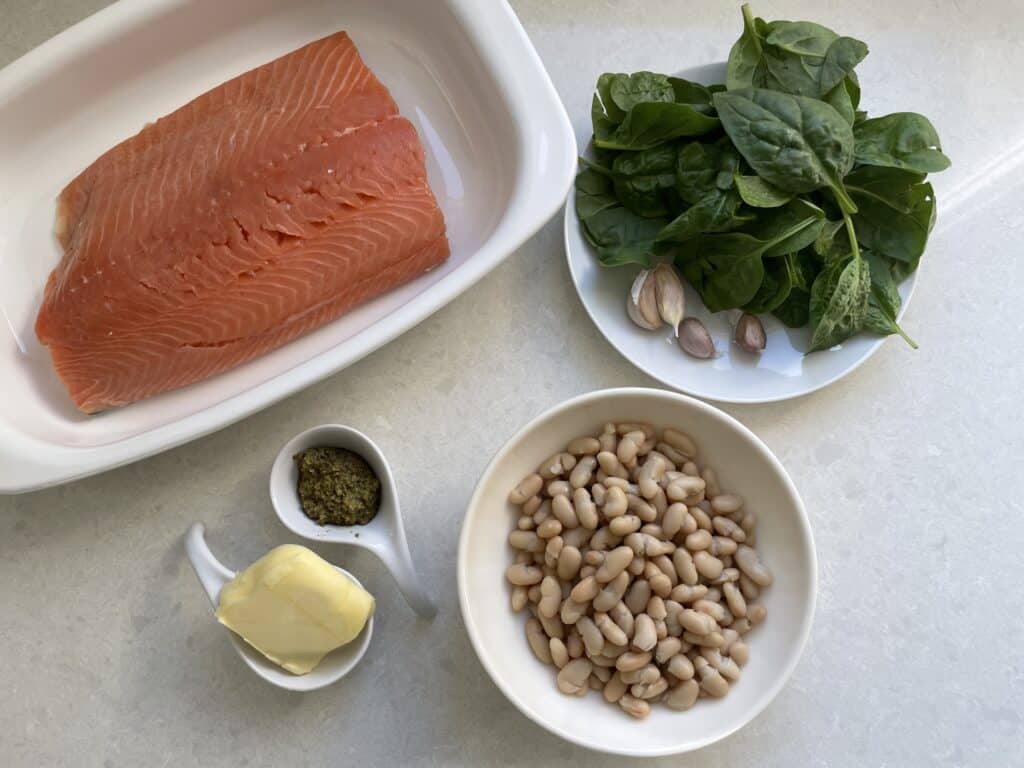 Ingredients in pesto butter salmon include salmon, spinach, garlic, pesto, butter and white kidney beans displayed on white countertop.
