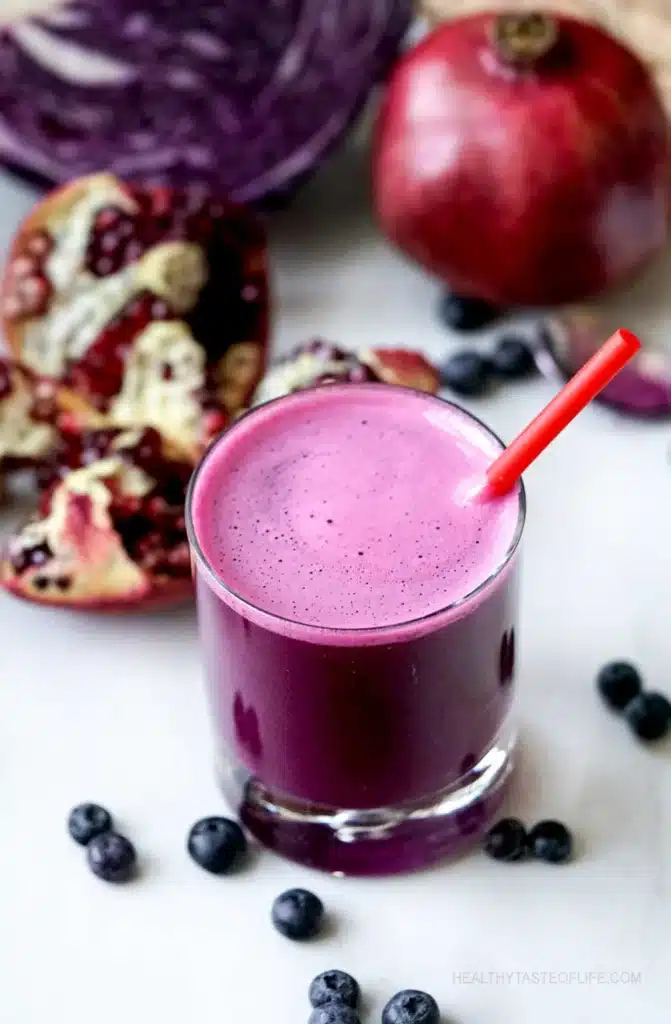 Purple juice to lower cholesterol presented in a clear glass with red straw surrounded by blueberries and an open pomegranate. 