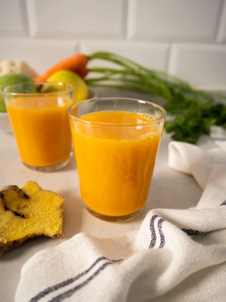 Two small clear orange juice to lower cholesterol on a white counter surrounded by carrots with the stem and ginger surround it.