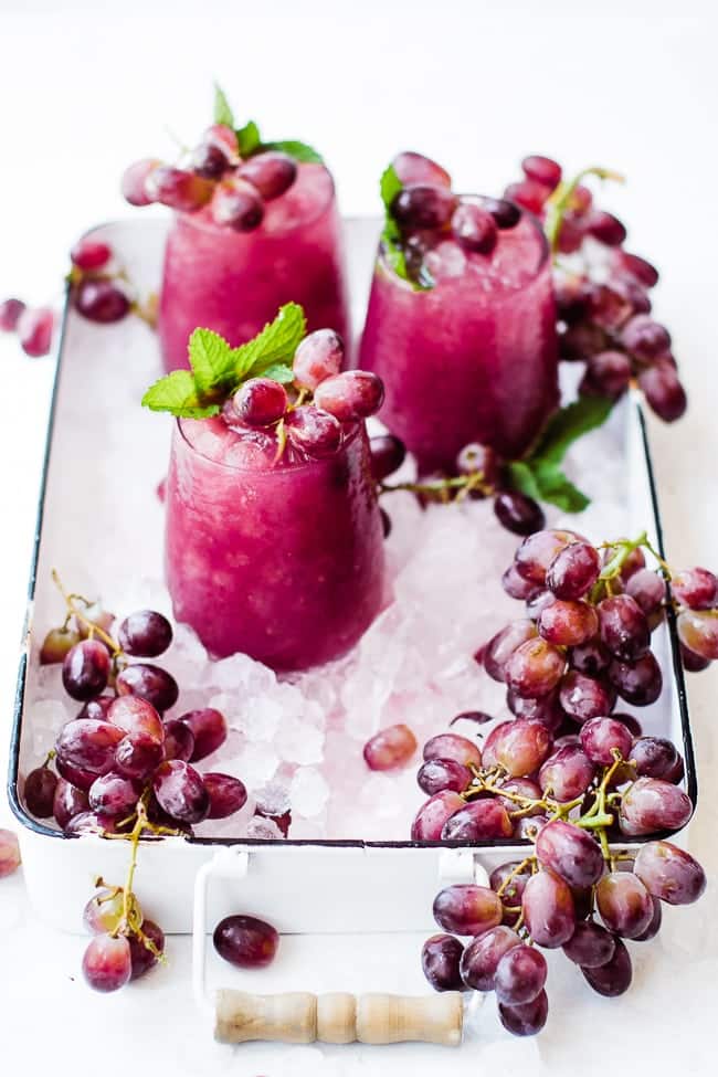 Two purple clear glasses on a tray with grapes topped with grapes and green herbs.