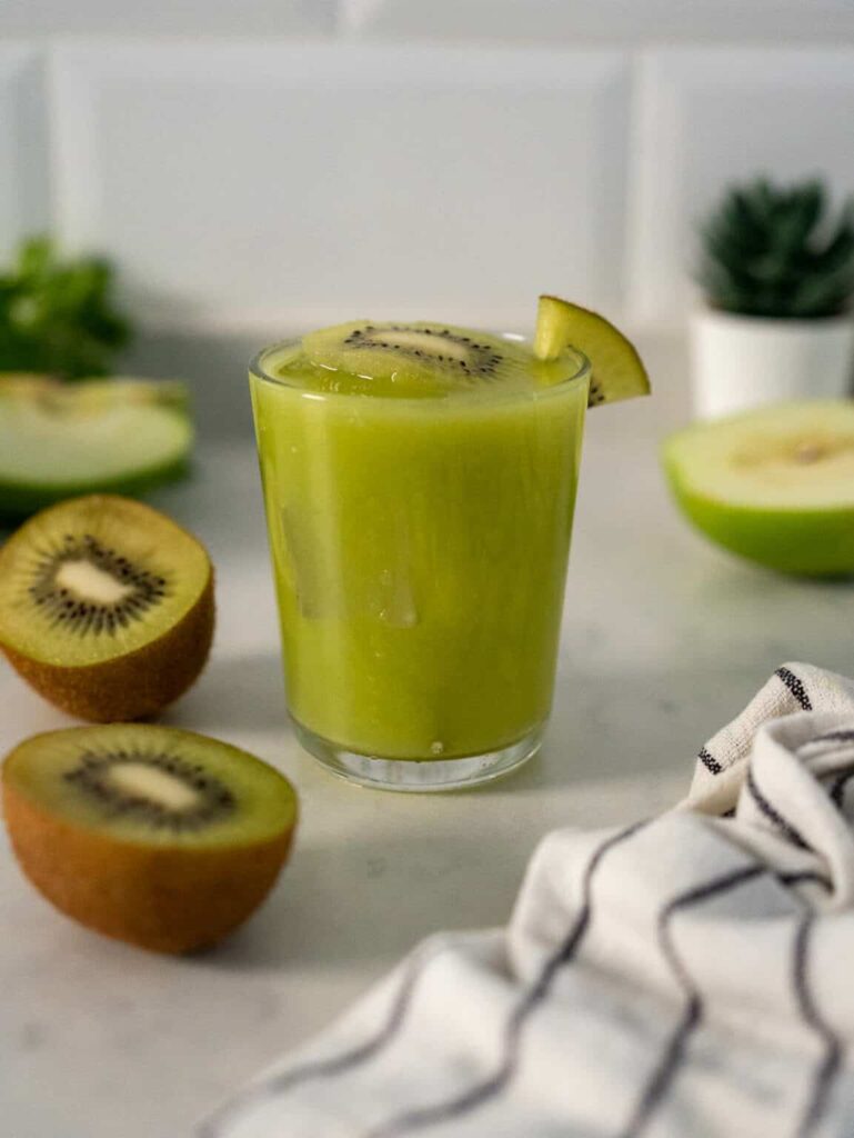 Short glass of green juice to lower cholesterol on a white counter in front of a white wall beside a kiwi cut in half.