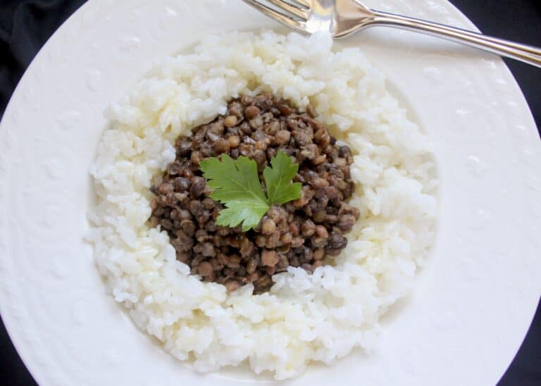 A picture of white plate with a mound of white rice topped with stewed green lentils with one single fresh herb on top.