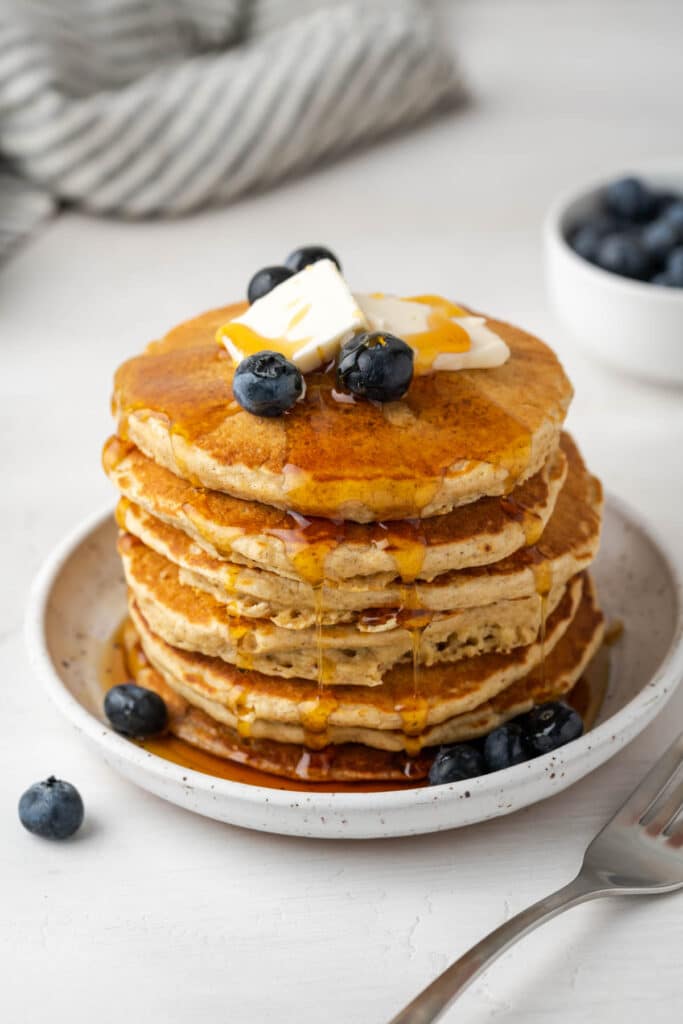 A stack of 10 pancakes topped with a square of butter and a few blueberries with amply syrup dipping down.