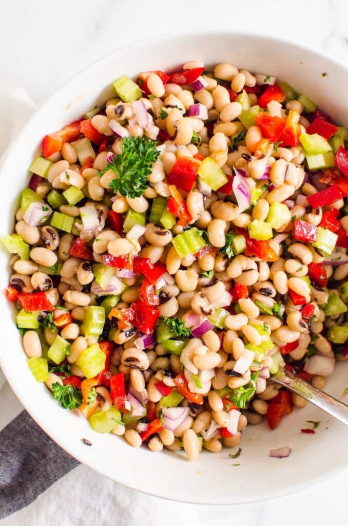 A large white bowl filled with white kidney beans, cubed peppers and celery topped with fresh herbs,