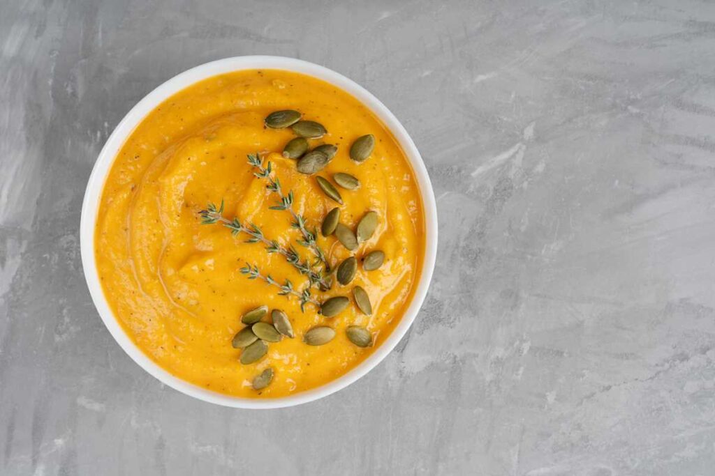 Picture of carrot lentil ginger soup in a white bowl on a grey background topped with pumpkin seeds,