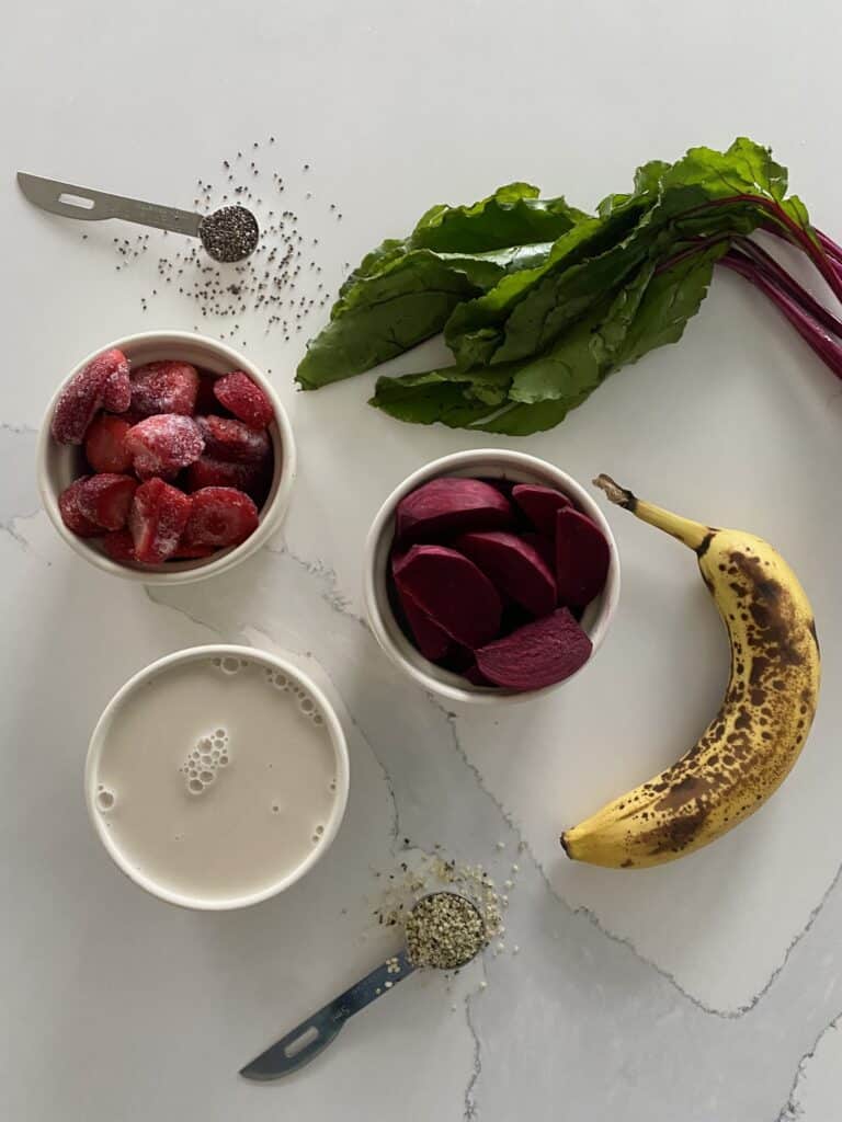 Beet greens smoothie ingredients on a white countertop beside three white bowls of soymilk, chopped beets, frozen strawberries, one banana, chia seeds, hemp seeds and beet greens on their stems.