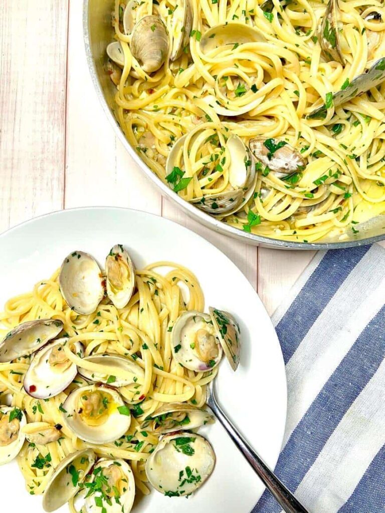 Creamy clam noodle dish presented in a skillet beside a plate of noodles with a fork, opened clam shells and topped with fresh herbs.