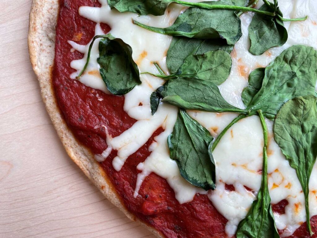 Low sodium pizza sauce on whole wheat pizza crust with melted cheese and cooked spinach on top presented on a wood cutting board.