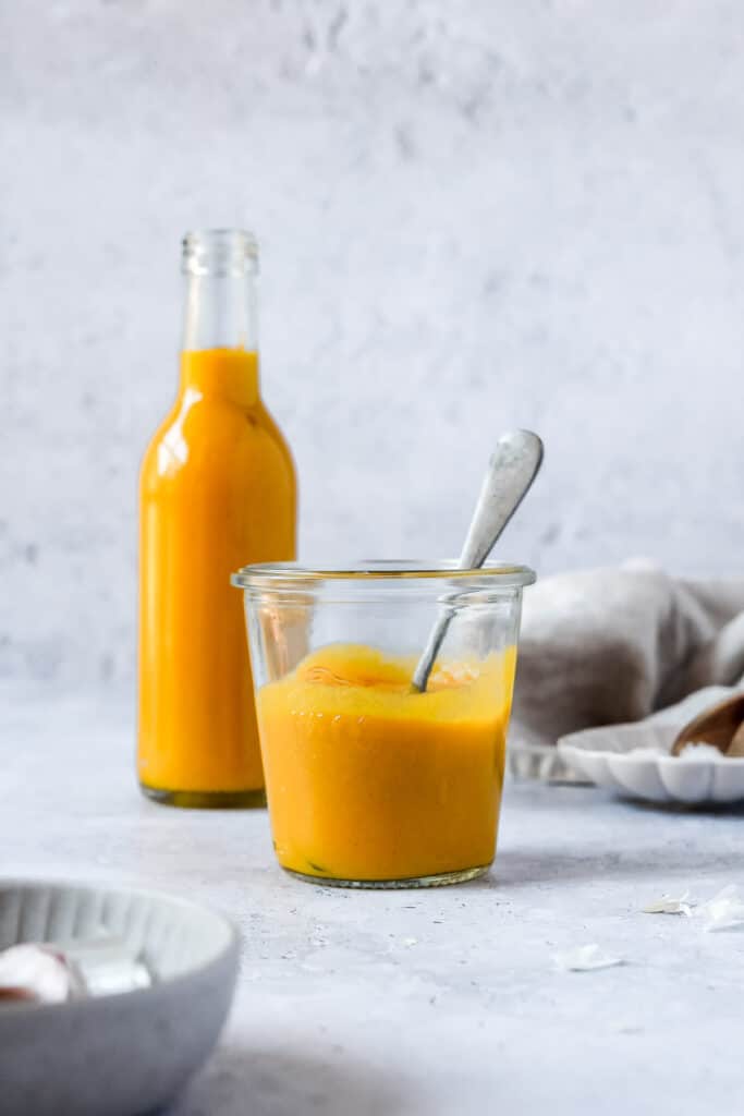 Mango sauce presented in a jar with a spoon in it beside another jar on a white counter with a white background beside a white tea towel.