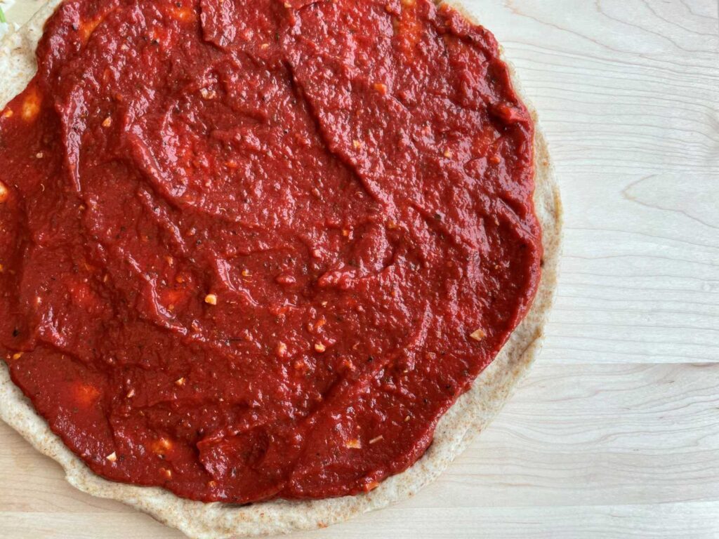 Low sodium pizza sauce on a whole wheat pizza crust on a wood cutting board.