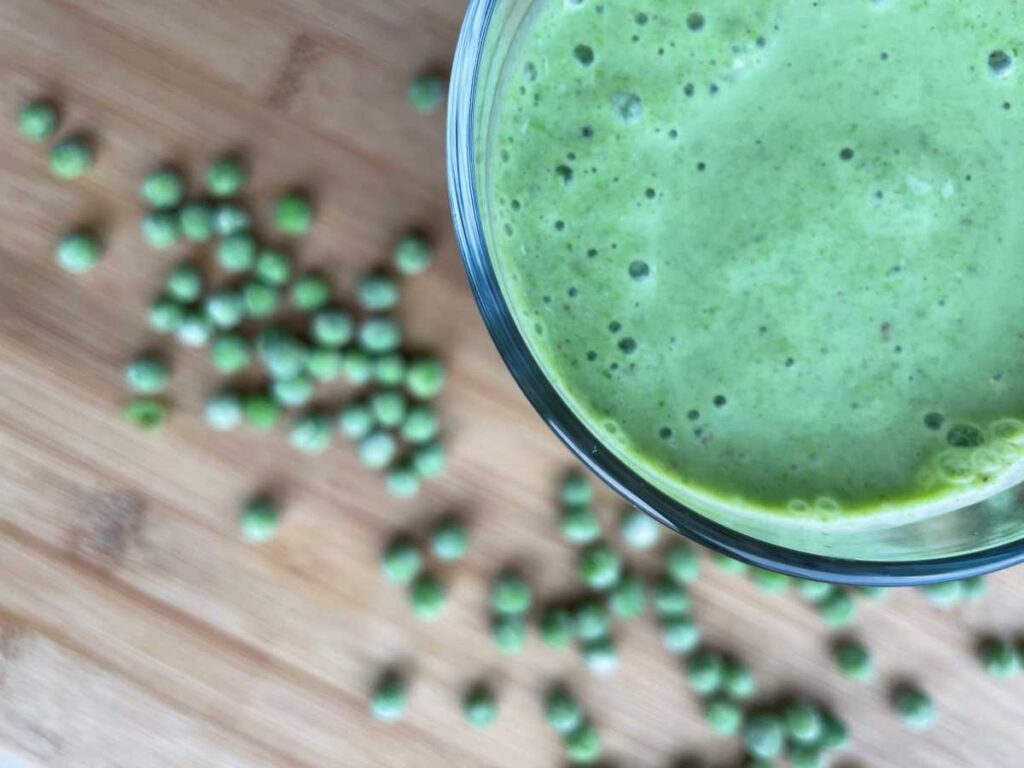 Green pea smoothie surrounded by frozen peas on a wood table.