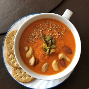 Protein tomato soup displayed in white mug bowl on white plate topped with pumpkin seeds, sesame seeds, cashews and a side of whole grain crackers.