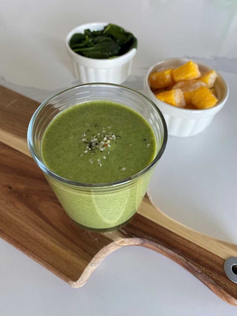 Green high potassium smoothie in a clear glass on a wood cutting board beside a white bowl of mango and a white bowl of frozen spinach.