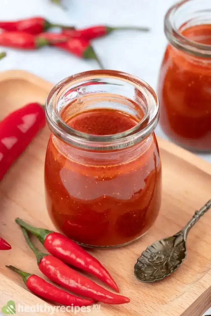 Red low sodium sauce in a clear jar beside another jar in the background surrounded by hot red and green peppers.