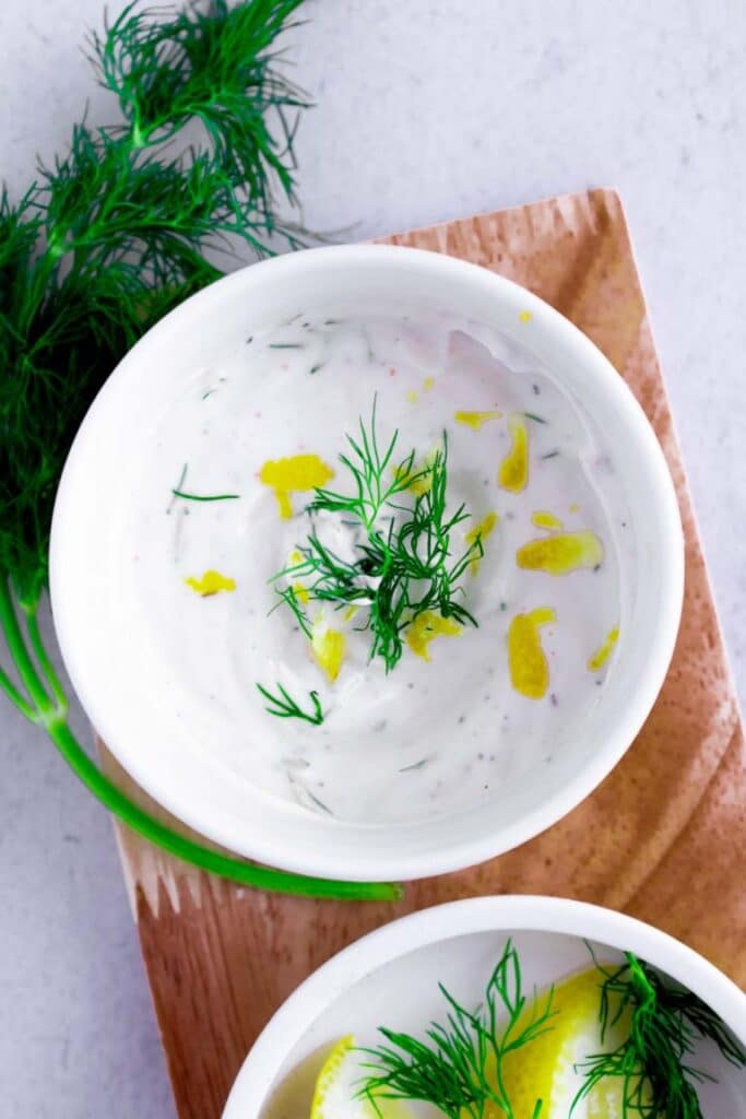 Low sodium sauce made of yogurt and dill presented in a white bowl on a wood board beside a bunch of dill.