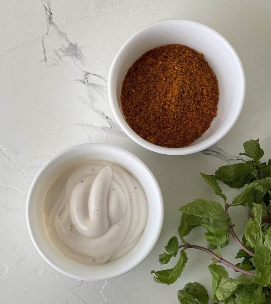 Two white bowls, one with mayo and the other red spices on a white counter beside a bunch of herbs.