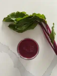 A white countertop with a glass of red beet smoothie on top from an aerial view. Beet stems are next to the smoothie glass.