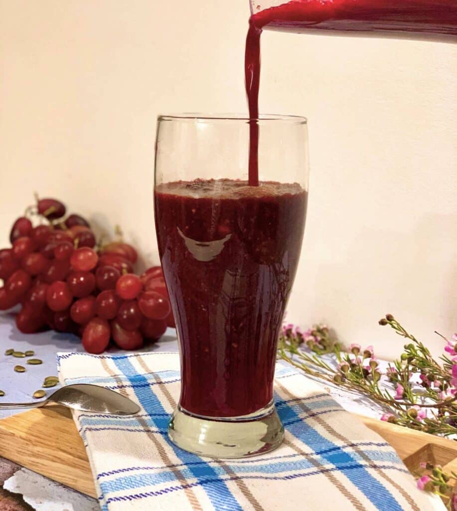 Image of a beet smoothie being poured into a glass on a table. A checkered tea towel is below the glass, with a bundle of grapes in the background.