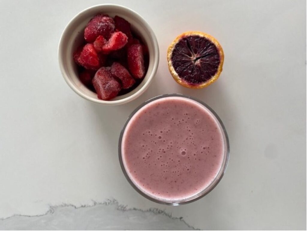 An aerial view of the calcium rich smoothie in a glass, on a white marble counter top. Next to the glass is a cup of sliced strawberries, and half a blood orange.