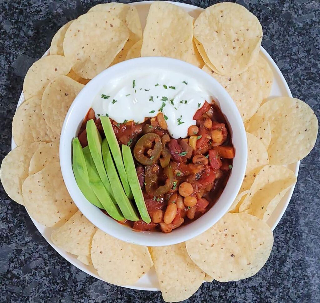 An aerial view of a bowl of chili on a plate surrounded by tortilla chips. The chili is topped with sliced avocado and sour cream.