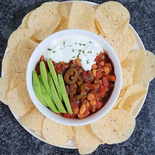 An aerial view of a bowl of chili on a plate surrounded by tortilla chips. The chili is topped with sliced avocado and sour cream.