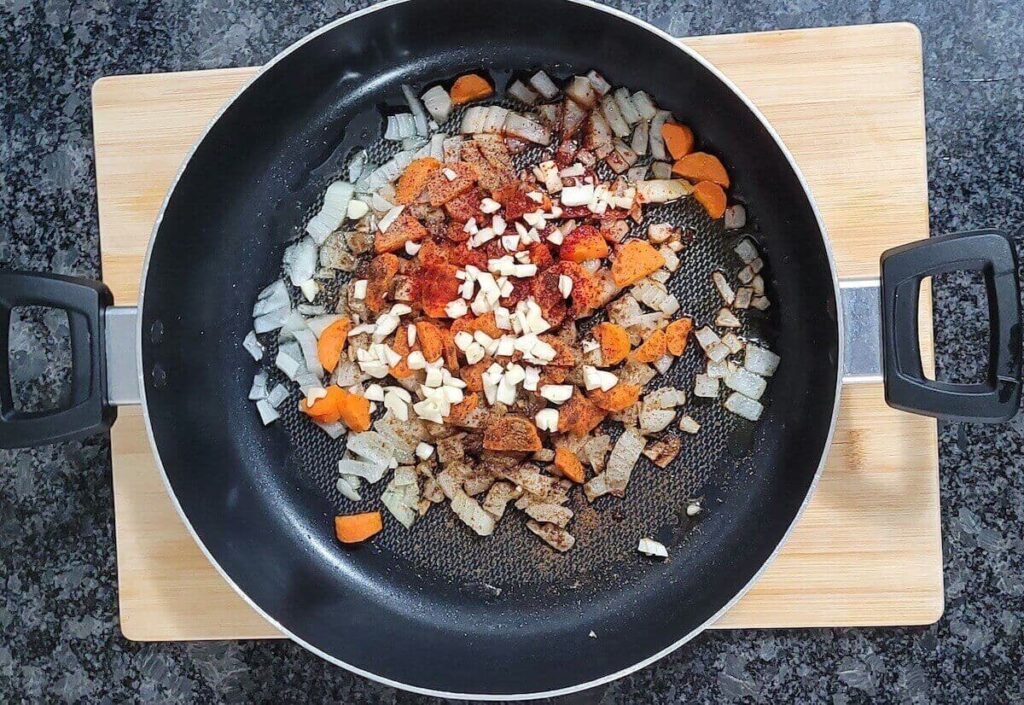 A pan placed on a cutting board resting on a grey and black counter top. In the pan is olive oil, carrot, onion, and spices before it is cooked. 