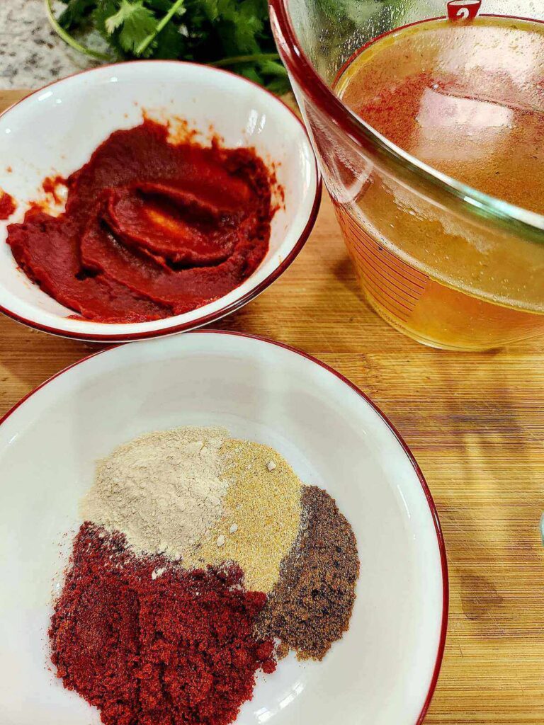 Low sodium enchilada sauce ingredients pictured. 3 containers are shown on a wooden cutting board. One with the spice mix, one with tomato paste, and the third contains vegetable broth.