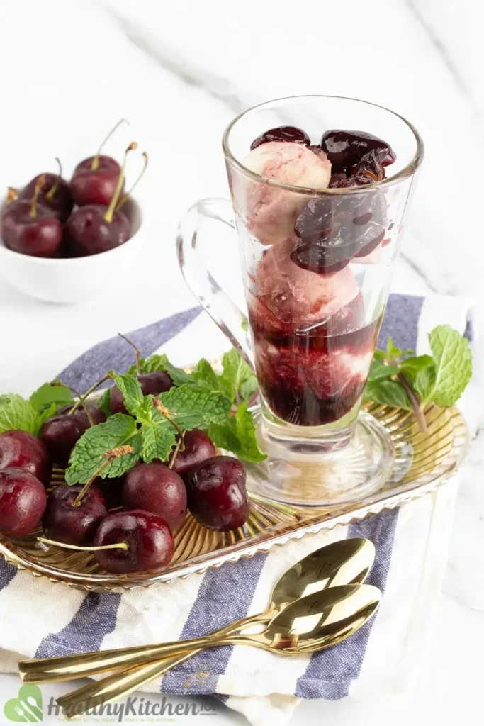 Yogurt topped with cherries beside a white bowl of cherries on a gold tray beside gold silverware.