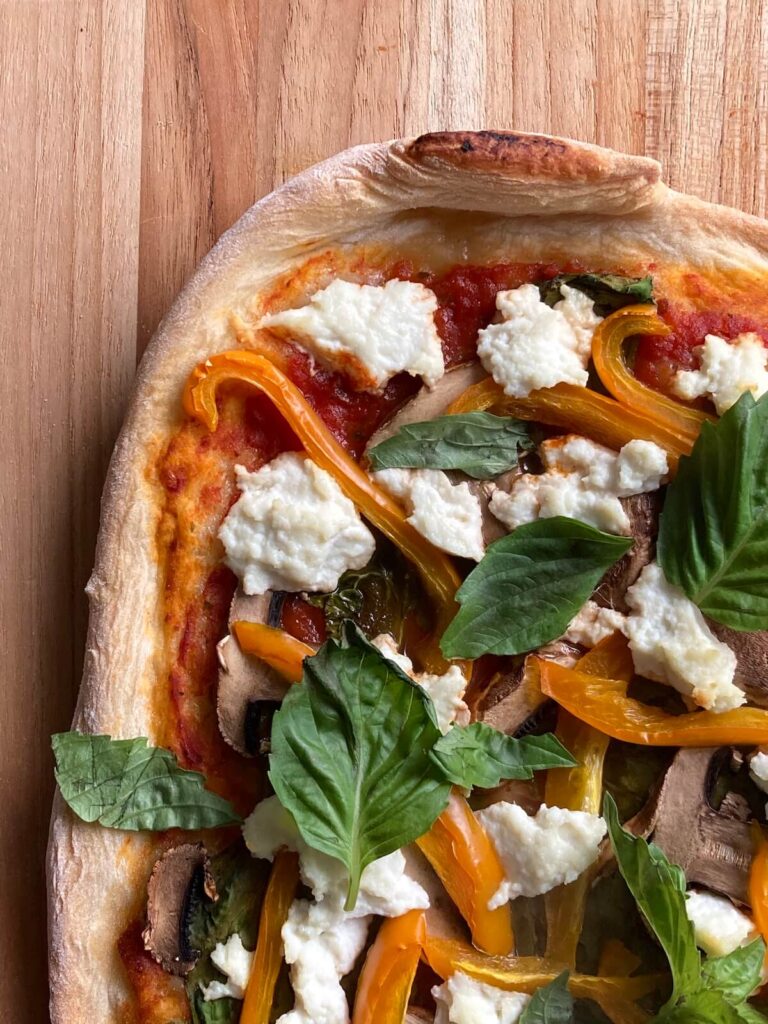 An image of pizza on top of a wooden cutting board. The pizza is topped with sauce, spinach, peppers, mushrooms, and cheese.