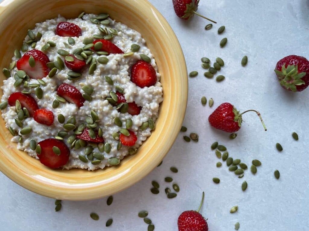 Mediterranean diet oatmeal in a yellow bowl, topped with strawberries and pumpkin seeds. Around the bowl are more scattered strawberries and pumpkin seeds.