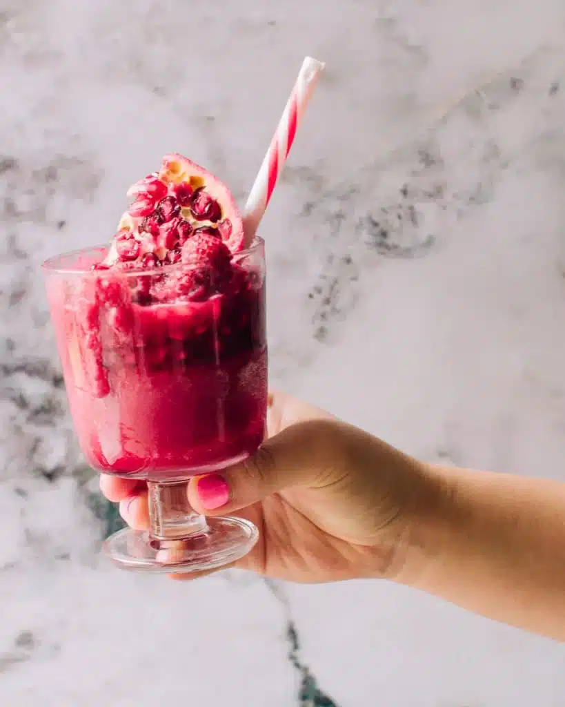 A pink beet and pomegranate smoothie in a glass with a straw, and piece of pomegranate on top. 