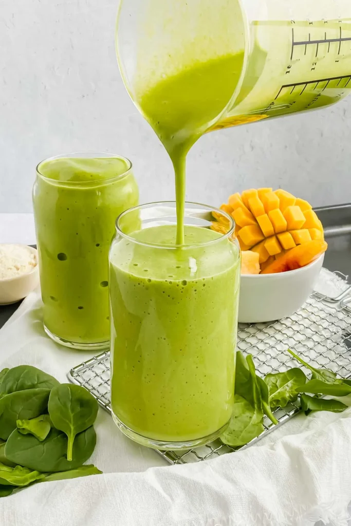 A green smoothie being poured into a glass. In the background is a bowl of Mano, and spinach. 