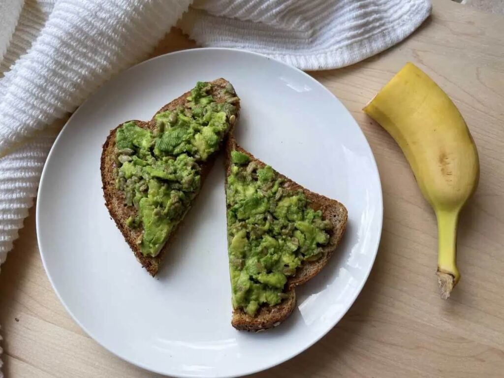 A slice of avocado peanut butter toast sliced in half. Half a banana is next to the plate.