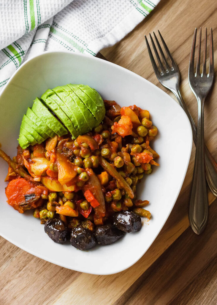Pictured is a vegetable stew topped with fresh avocado, with two forks next to the dish. 