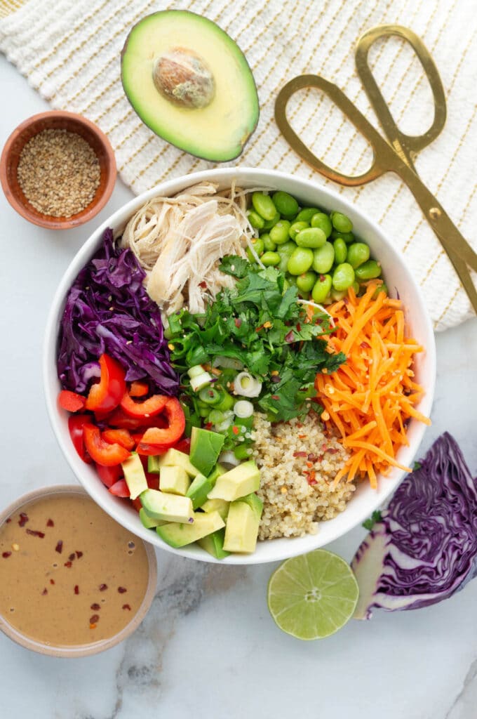 Thai quinoa salad pictured in a white bowl, with peanut sauce, cabbage, lime, and avocado surrounding the bowl.