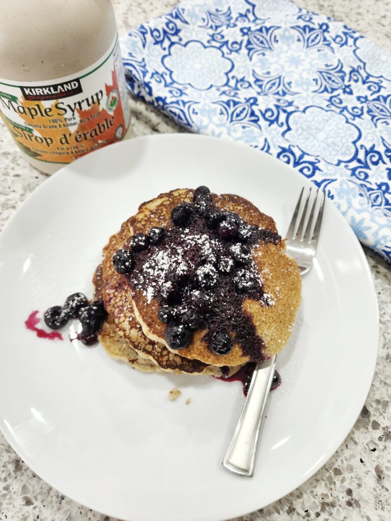 Barley pancakes stacked on white plate beside a fork, topped with blueberries and sugar beside a bottle of maple syrup and a blue and white napkin.