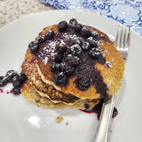 Barley pancakes with a fork stacked on white plate, topped with blueberries and sugar beside and a blue and white napkin.